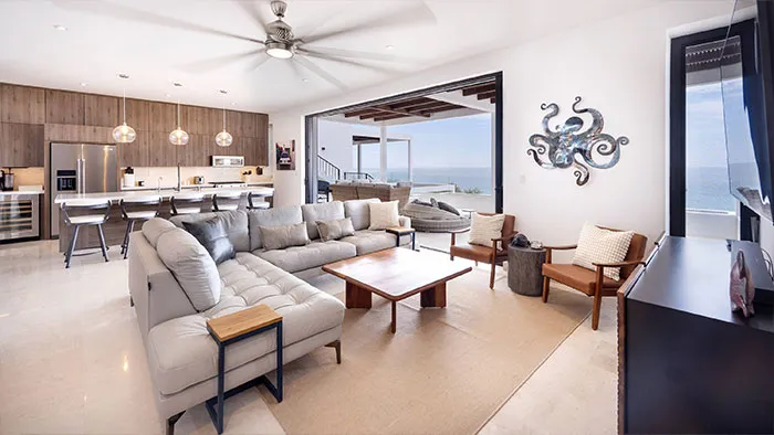 The Cove At Cerritos 402 Living Room - Spacious living room in Residence 402 with a large sectional sofa, modern armchairs, and wall-mounted octopus art, opening to an ocean-view terrace through wide sliding glass doors.